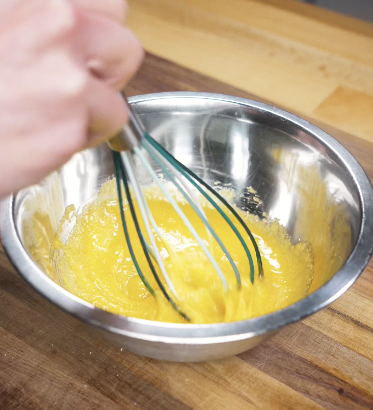 Egg yolks and sugar are whisked together in a metal mixing bowl, forming the rich yellow base of the tiramisu cream.