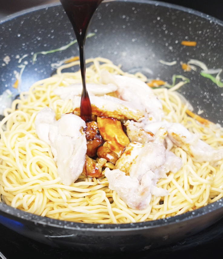 Dark savory sauce being poured over cooked chicken and noodles in a wok, capturing the beginning of flavor infusion.