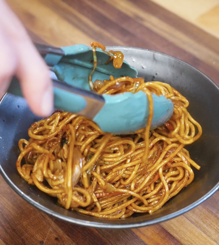 Blue tongs scooping a portion of stir-fried chicken noodles into a bowl, highlighting the glossy, saucy texture.