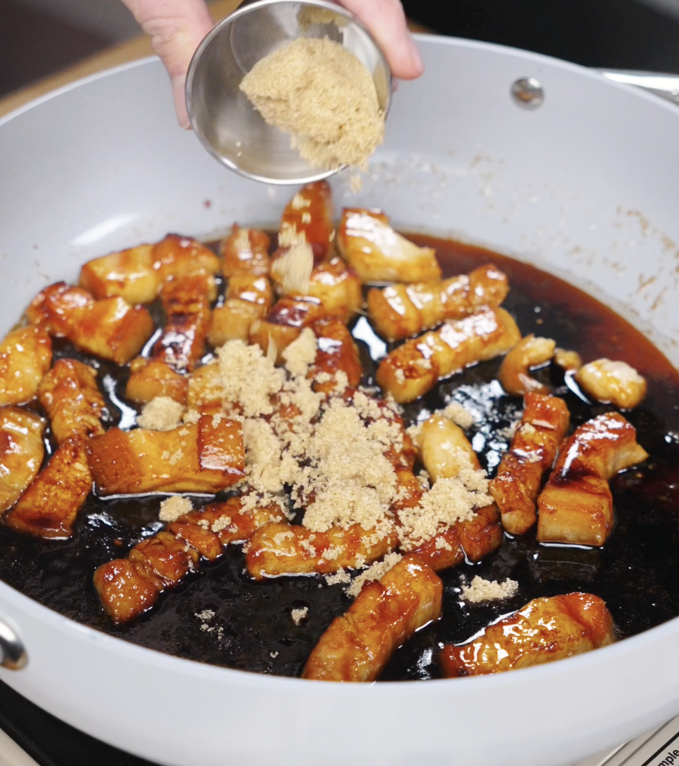 A hand sprinkling brown sugar over glazed pork belly in a skillet filled with soy sauce, creating a rich caramel base.
