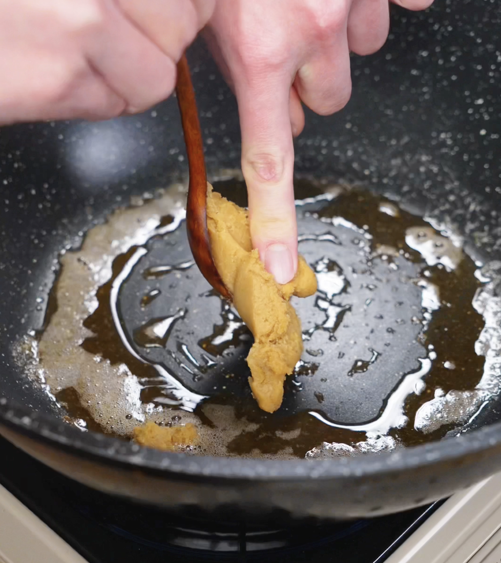 A hand presses miso paste off a wooden spoon into a pan of browned butter, beginning the rich sauce for the noodles.