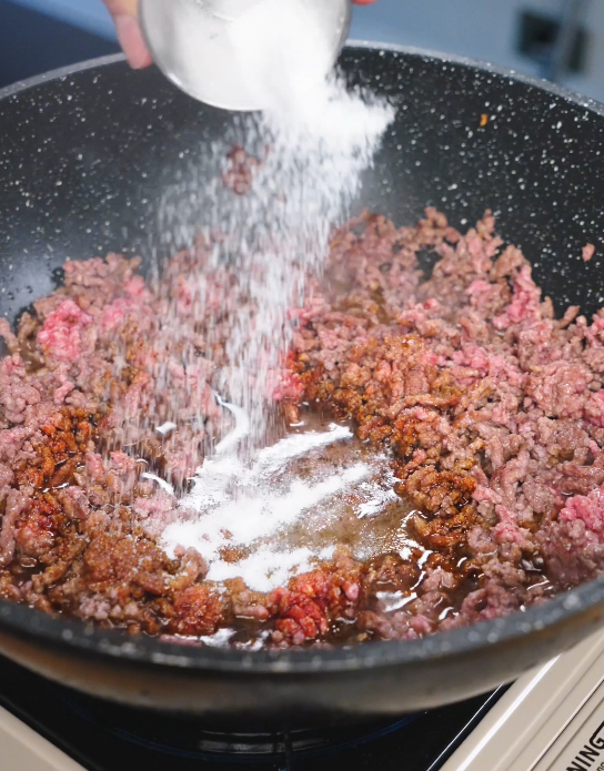 Sugar being sprinkled into the pan with browned ground beef and sauce, helping create the signature sweet and savory Mongolian glaze.