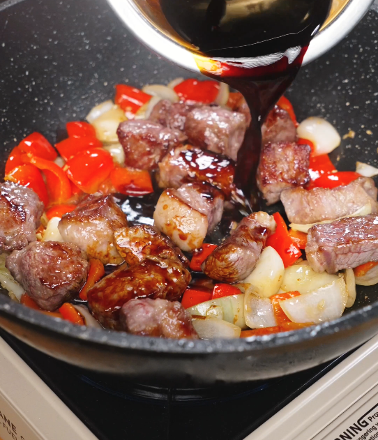 Black pepper sauce being poured over seared beef and vegetables in the pan, beginning to simmer and coat the ingredients.