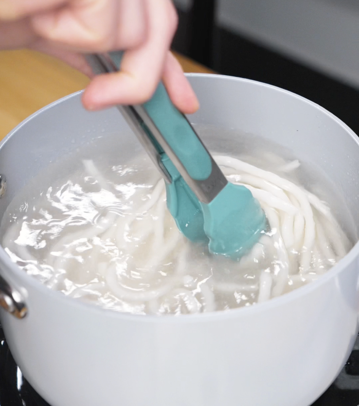Tongs stirring thick udon noodles in a pot of boiling water, just before they reach al dente.