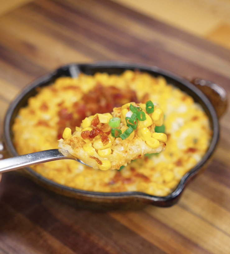 A spoonful of hot, melty Korean corn cheese lifted from the pan, showing corn, gooey cheese, and scallion topping.