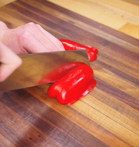 Red bell pepper being sliced into chunky pieces on a wooden cutting board, prepped for black pepper beef.