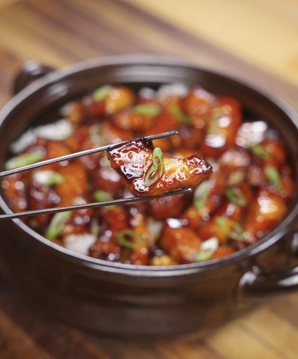 A glossy piece of pork belly lifted by chopsticks, showcasing the thick caramel glaze and a garnish of scallion.