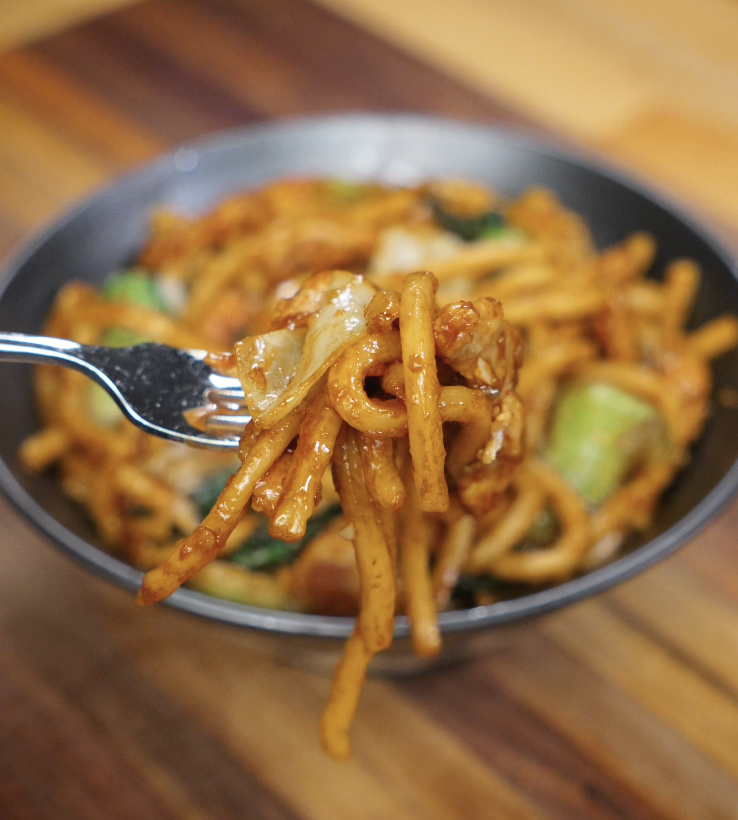 A fork lifting saucy udon noodles coated in glossy soy sauce with cabbage and chicken visible in the background.