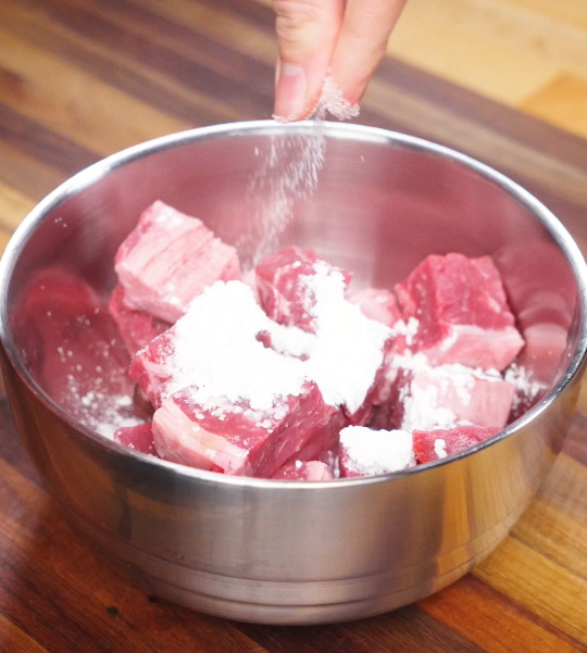 Cubed beef tossed with cornstarch and seasoning in a mixing bowl, creating a light coating that helps lock in moisture during cooking.