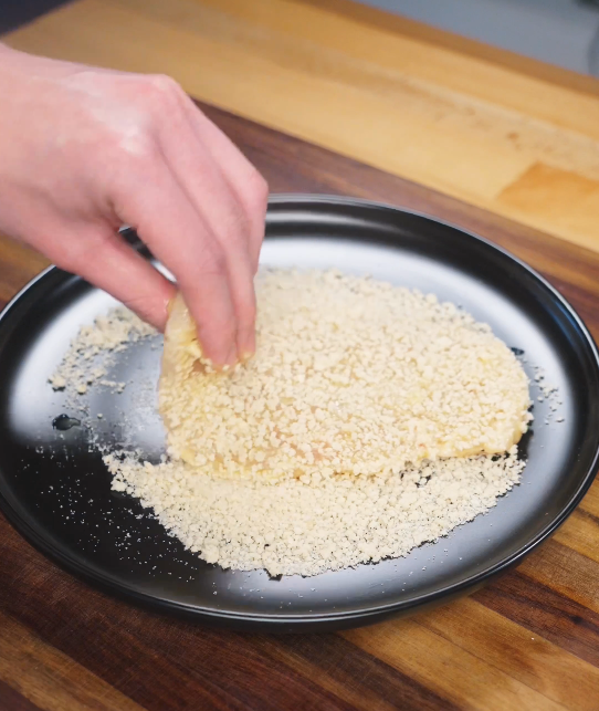 Thinly sliced chicken breast being pressed into panko breadcrumbs on a plate, fully coating the surface to create a crispy air fryer chicken katsu crust.