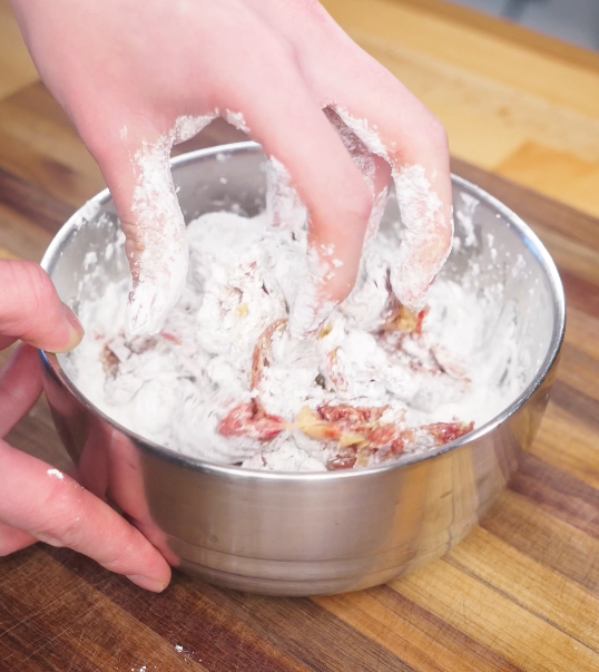 Thin slices of marinated beef being coated in cornstarch by hand in a metal bowl, creating a crisp layer before frying.