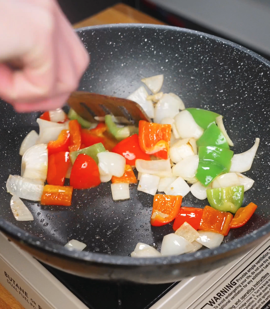 Onion wedges and red and green bell peppers being stir-fried in a pan until slightly softened and fragrant.