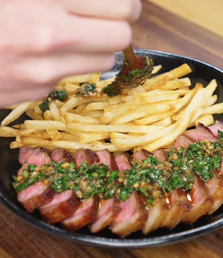 A spoon pouring fresh chimichurri over sliced steak and crispy fries, letting the herbs and oil soak into each piece for maximum flavor.