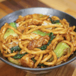 A serving bowl filled with glossy Shanghai Fried Udon Noodles, stir-fried with tender chicken, bok choy, and cabbage in a rich soy-based sauce.