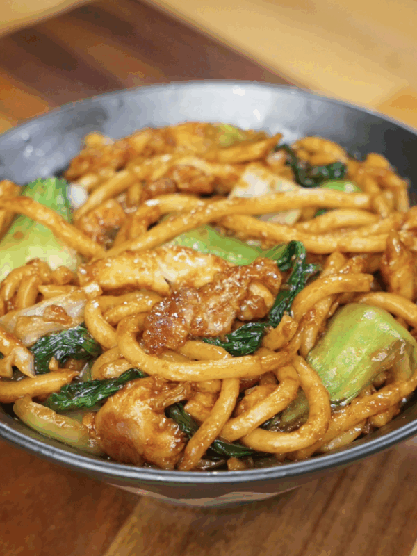 A serving bowl filled with glossy Shanghai Fried Udon Noodles, stir-fried with tender chicken, bok choy, and cabbage in a rich soy-based sauce.