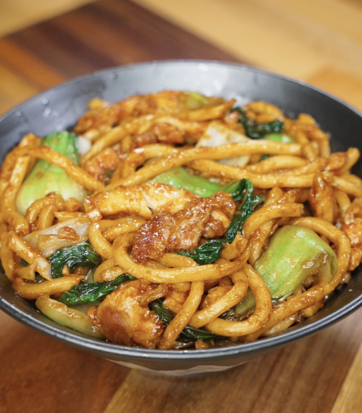 A serving bowl filled with glossy Shanghai Fried Udon Noodles, stir-fried with tender chicken, bok choy, and cabbage in a rich soy-based sauce.