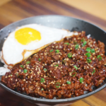 A bowl of Mongolian ground beef served over white rice with a crispy fried egg, garnished with sesame seeds and scallions.