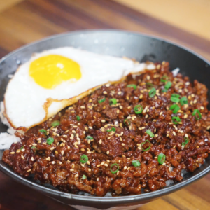 A bowl of Mongolian ground beef served over white rice with a crispy fried egg, garnished with sesame seeds and scallions.