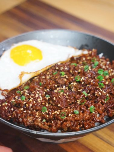 A bowl of Mongolian ground beef served over white rice with a crispy fried egg, garnished with sesame seeds and scallions.