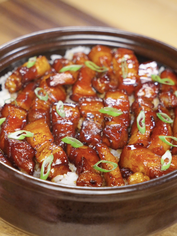 A ceramic bowl filled with caramelized pork belly cubes glazed in a dark sticky sauce, served over white rice and garnished with sliced scallions.