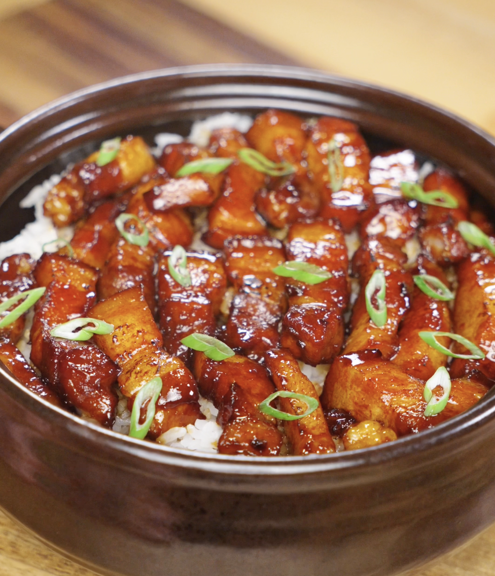 A ceramic bowl filled with caramelized pork belly cubes glazed in a dark sticky sauce, served over white rice and garnished with sliced scallions.