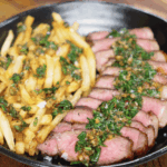 A cozy plate of steak frites with crispy golden fries and perfectly pink sliced steak, topped with fresh chimichurri for a vibrant, herb-packed finish.