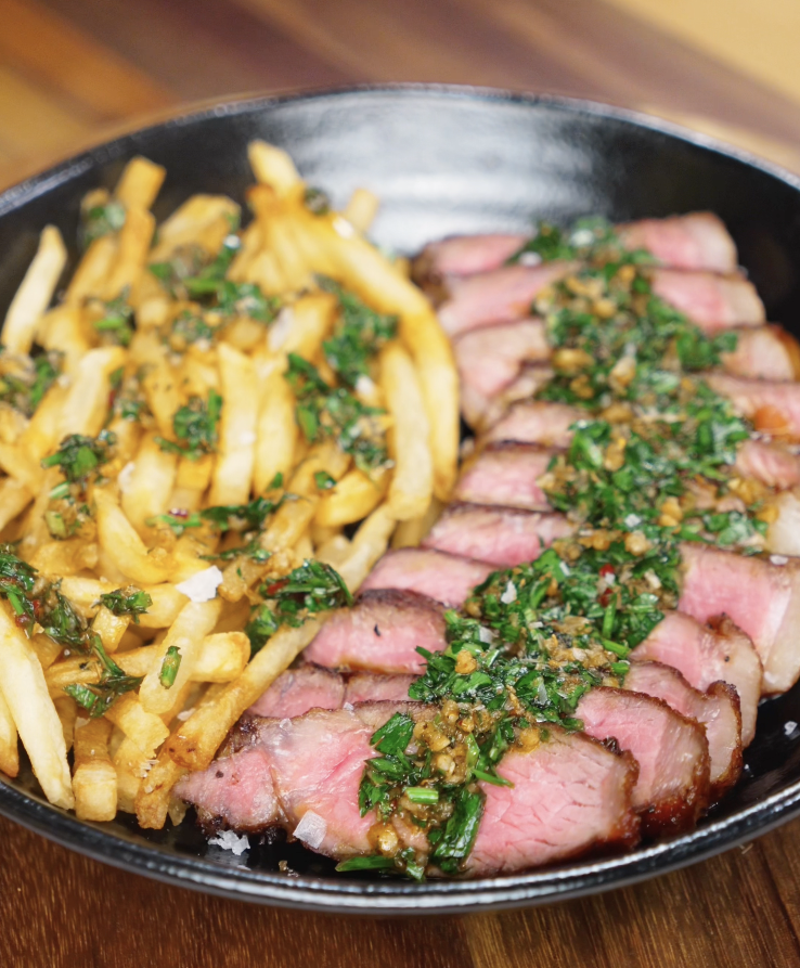 A cozy plate of steak frites with crispy golden fries and perfectly pink sliced steak, topped with fresh chimichurri for a vibrant, herb-packed finish.