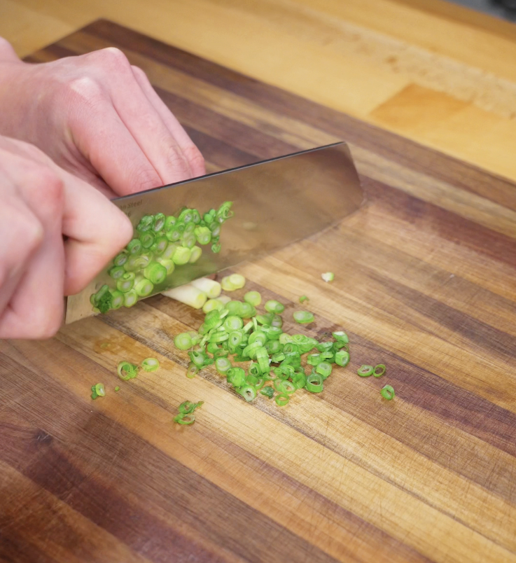 Close-up of hands finely chopping scallions on a wooden cutting board, preparing garnish for Korean corn cheese.