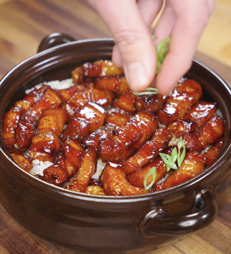 A hand adding freshly sliced scallions on top of caramelized pork belly in a serving bowl for a bright finishing touch.