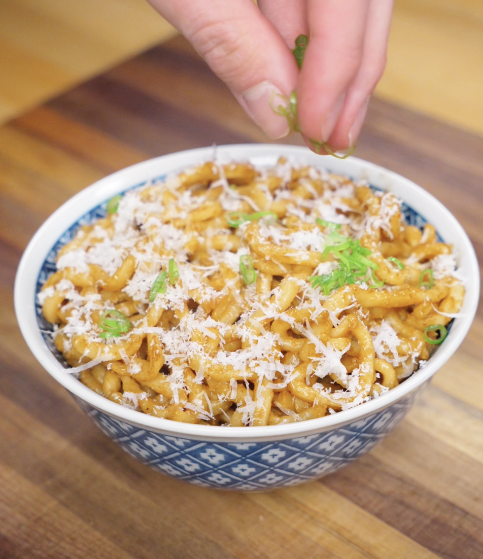 A hand sprinkles sliced scallions over a bowl of brown butter miso noodles topped with grated parmesan for a fresh finish.