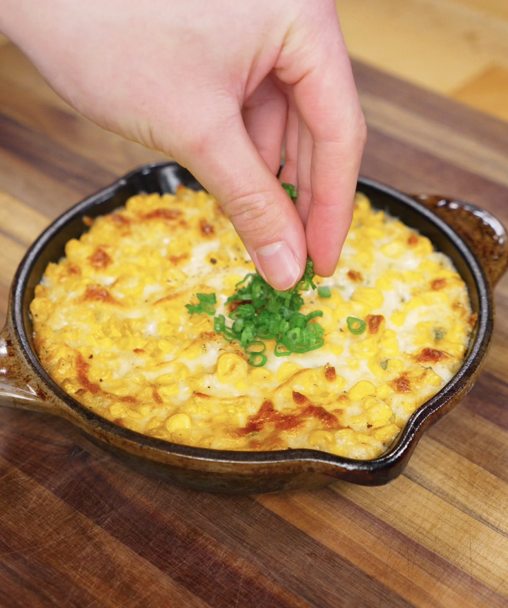 A hand sprinkling sliced scallion greens over the hot, bubbly Korean corn cheese for a fresh pop of color and flavor.
