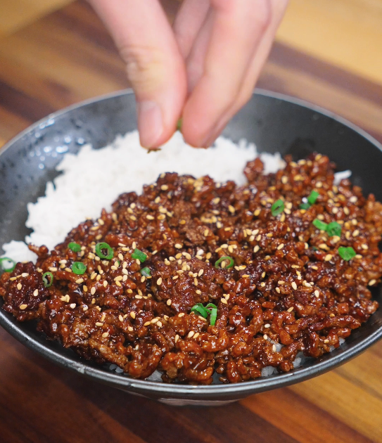 Fresh scallions and sesame seeds being sprinkled over Mongolian ground beef, adding color and texture before serving.