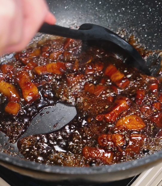 Pork belly being stirred through a thickened, bubbling sauce as the glaze clings to the meat, creating a rich, lacquered finish.