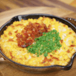 Finished Korean corn cheese topped with crispy bacon bits and scallion greens, ready to serve in a cast iron dish.
