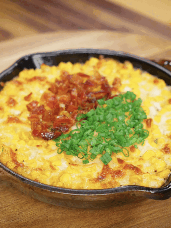Finished Korean corn cheese topped with crispy bacon bits and scallion greens, ready to serve in a cast iron dish.