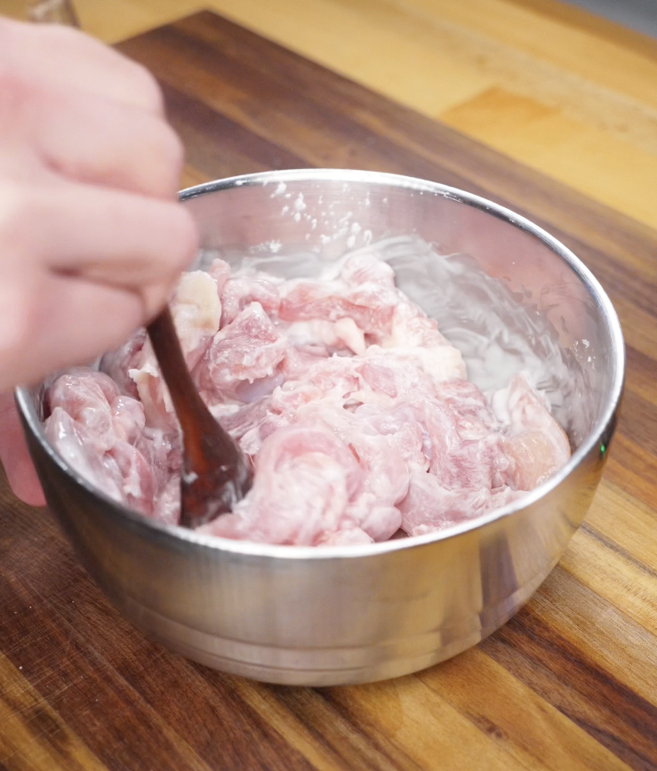 Raw sliced chicken thighs being mixed with shaoxing wine, cornstarch, and salt in a silver mixing bowl.
