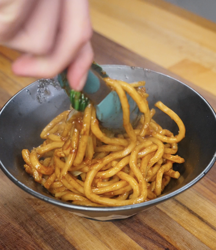 A hand uses tongs to lift a serving of saucy Shanghai fried noodles into a dark bowl, steam rising from the glossy noodles coated in savory sauce.