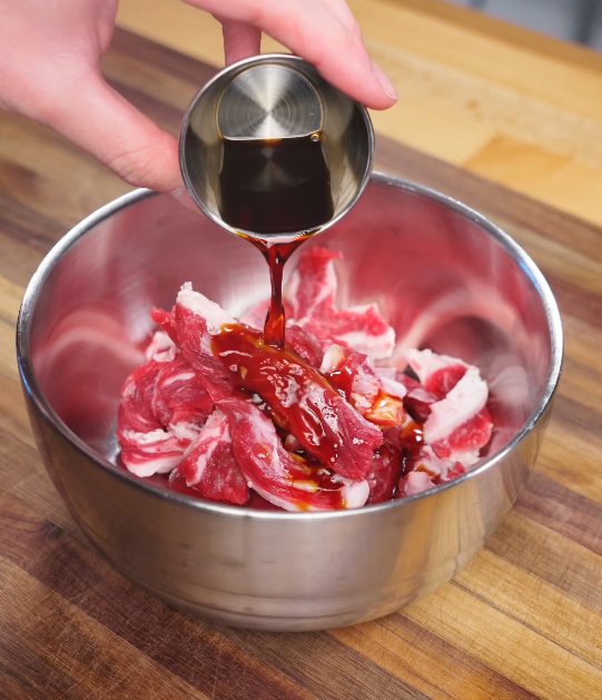 Soy-based marinade being poured over raw sliced beef in a metal bowl, beginning the flavor-building step of Beijing Beef.