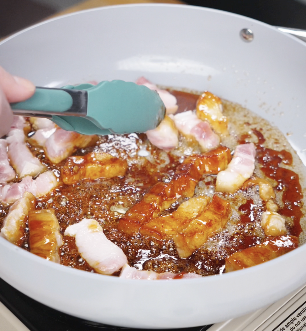 Raw cubes of pork belly cooking in bubbling caramel, some pieces beginning to brown while being flipped with tongs.