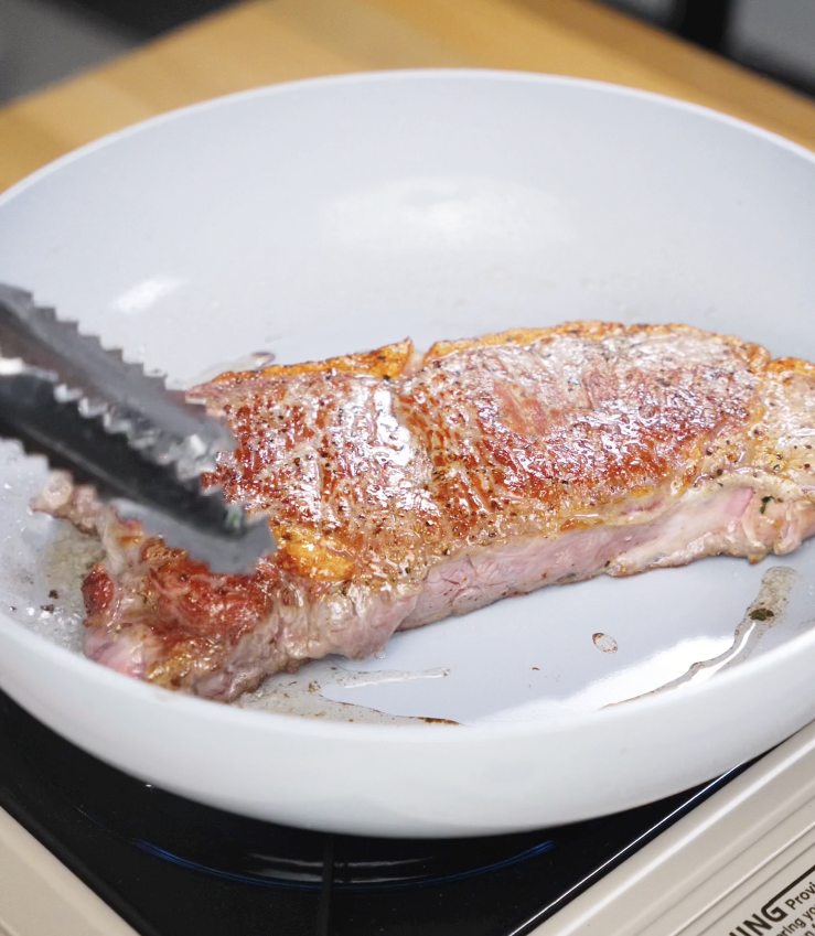 A thick-cut steak sizzling in a hot pan, developing a deep golden-brown crust as it sears to lock in juices and build rich flavor.