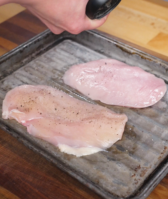 Raw chicken breasts laid out on a baking tray being seasoned with salt and black pepper before dredging for chicken katsu.