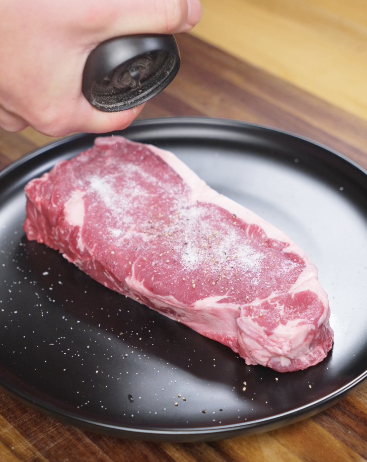 A raw NY strip steak being generously seasoned with salt and pepper on a dark plate, prepping it for a perfect sear.