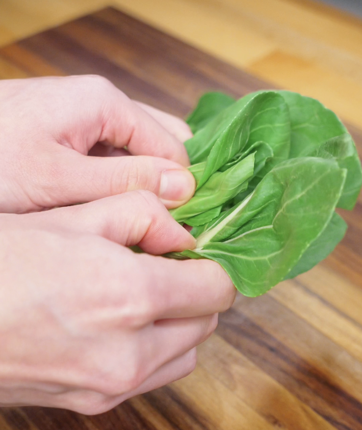 Hands gently separating baby bok choy leaves on a wooden cutting board for stir-frying.