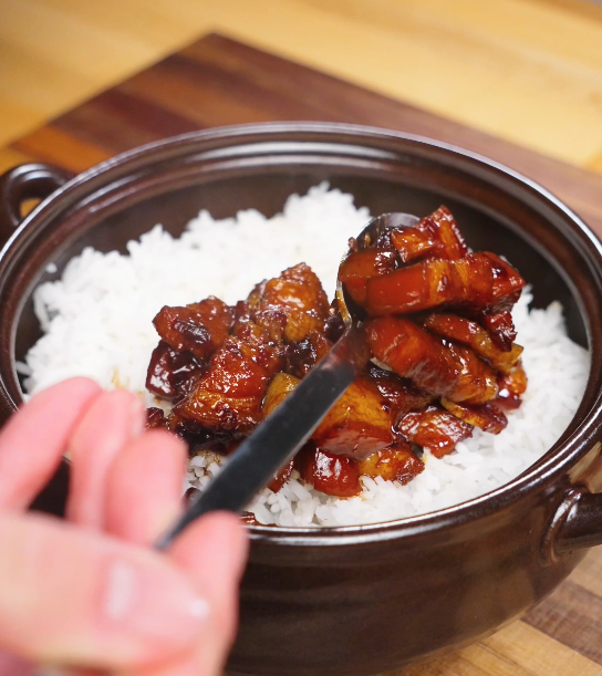 Spoonfuls of red braised pork belly being placed over freshly steamed white rice, with the glossy sauce soaking into the grains.