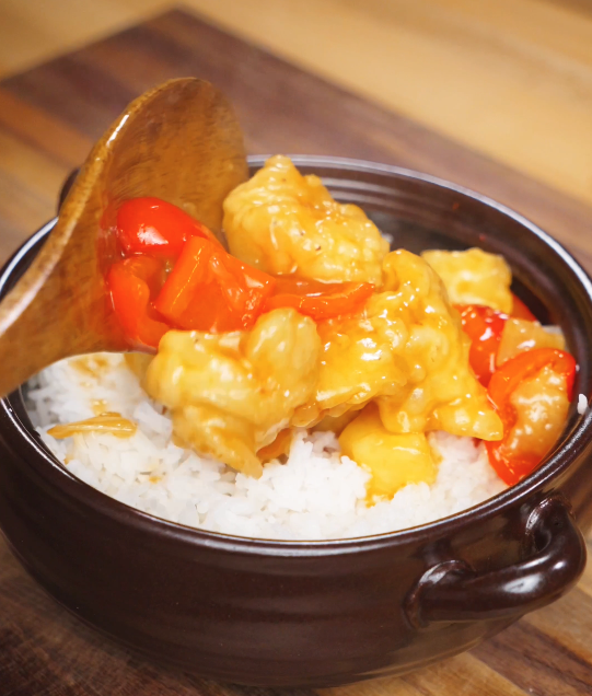 Sweet and sour chicken being spooned over a bowl of steamed white rice, with glossy battered chicken, bell peppers, and pineapple coated in a vibrant tangy sauce.