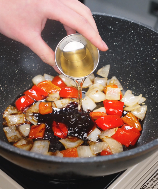 Chopped onions and red bell peppers sizzling in a wok as a hand pours vinegar into the glossy Beijing Beef sauce base.