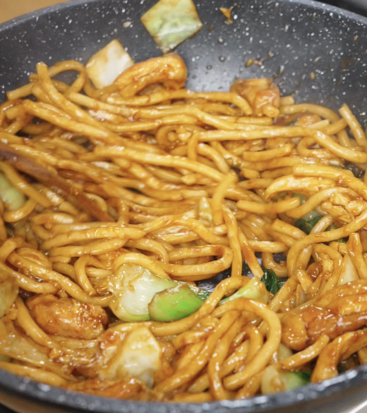 A close-up of thick udon noodles being tossed in a hot pan with tender chicken, cabbage, and a glossy soy-based sauce for Shanghai fried noodles.