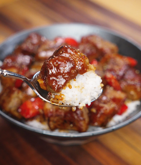 A spoonful of black pepper beef lifted over steamed white rice, showing tender beef cubes coated in a glossy, pepper-forward sauce.