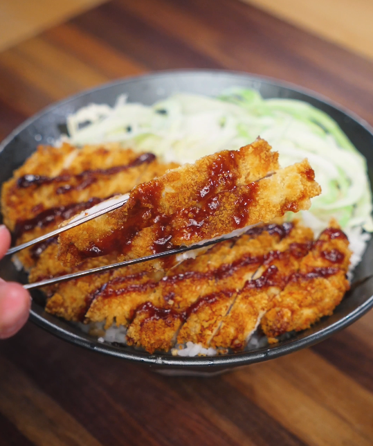 Sliced air fryer chicken katsu lifted with chopsticks over a bowl of rice, showing the crunchy golden coating and juicy interior with tonkatsu sauce drizzled on top.
