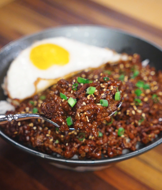 A spoonful of glossy Mongolian ground beef lifted from the bowl, showing the sticky sauce clinging to the beef with rice underneath.